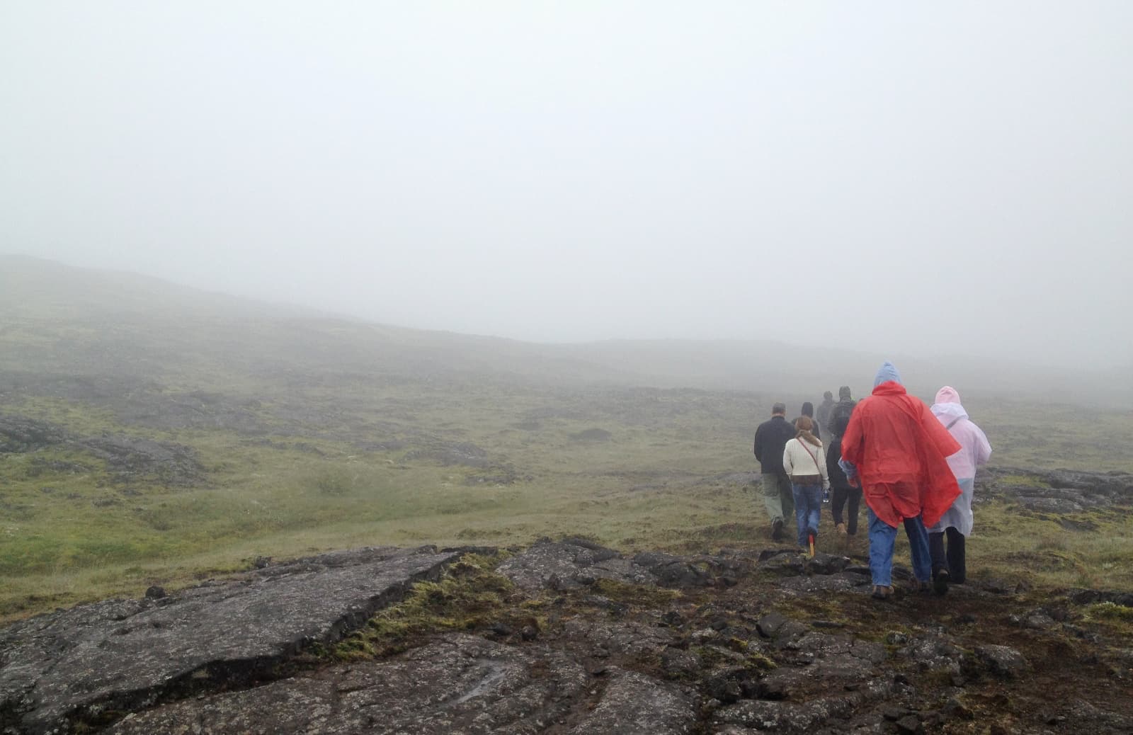 A cluster of people walking across rugged rocky and mossy terrain, with hazy moist skies in the distance